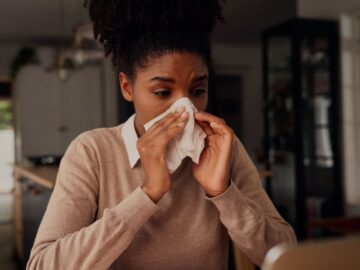 A woman in a tan sweater sits at a table, holding a white tissue up to her nose. The background is blurry.