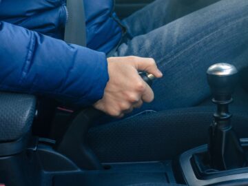 A person wearing a puffy, blue jacket pulls the parking brake in a car. They are wearing their seatbelt.