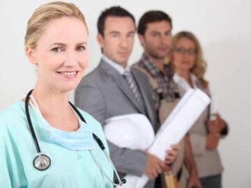 A female nurse wearing scrubs and a stethoscope standing in front of three other professionals from different industries.