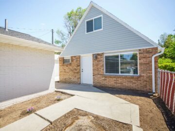 A sidewalk leads to the front door of an accessory dwelling unit made of bricks and blue vinyl siding. The ADU is behind a house.