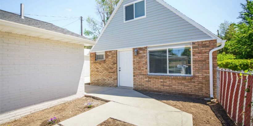 A sidewalk leads to the front door of an accessory dwelling unit made of bricks and blue vinyl siding. The ADU is behind a house.