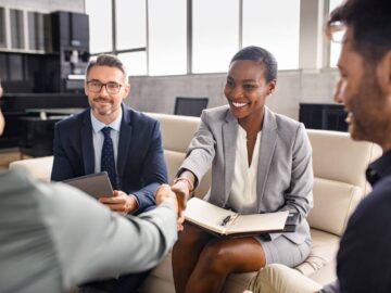 A business woman sitting on a couch with her colleagues as she smiles and shakes the hand of a client.