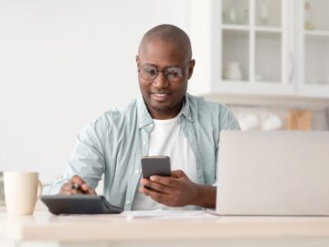 Black male investor sitting down at the table with his phone, a tablet, and a laptop. He's taking notes about his portfolio.