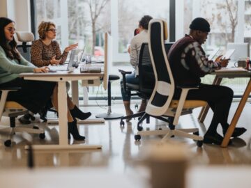 Employees with laptops or tablets sit at desks in an open office space near floor-length windows, as two teammates chat.