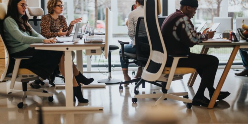 Employees with laptops or tablets sit at desks in an open office space near floor-length windows, as two teammates chat.