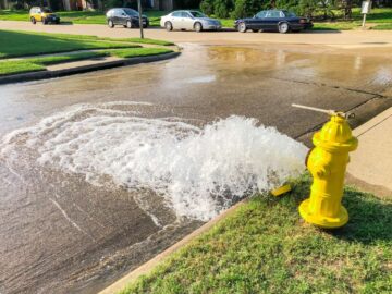 A yellow fire hydrant gushes water onto a residential street. A wrench is attached to the top of the hydrant.