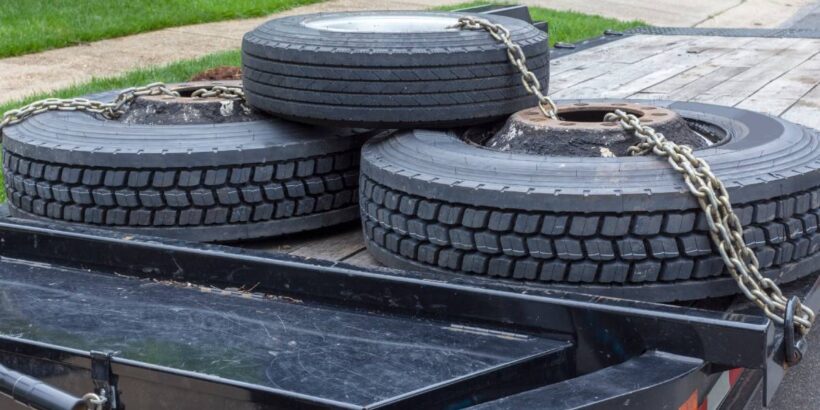 A flatbed trailer carrying three large tires secured with heavy chains, parked on a suburban street near a grassy lawn.