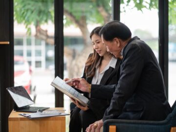 A man and woman dressed in business clothing reviewing documents and data on a laptop in an office with large windows.