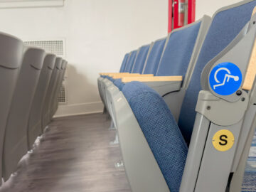 A close-up of an auditorium row of blue seats with one marked as for wheelchair users on the side of the grey armrest.