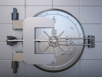 A circular vault door is closed against the surrounding metal wall. The door has large hinges and a four-spoke handle.