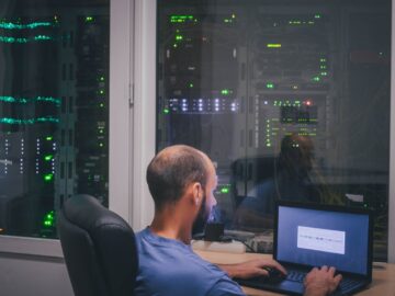 A programmer sits at a desk outside a data center with glass partition walls. Lights are visible in the dark room.