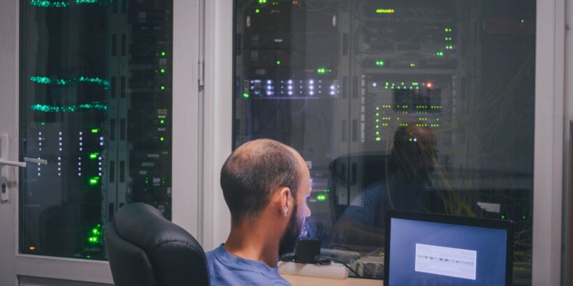 A programmer sits at a desk outside a data center with glass partition walls. Lights are visible in the dark room.