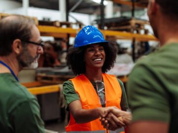 A black woman wearing a blue hard hat and orange vest shakes hands with someone. Another man stands next to them.