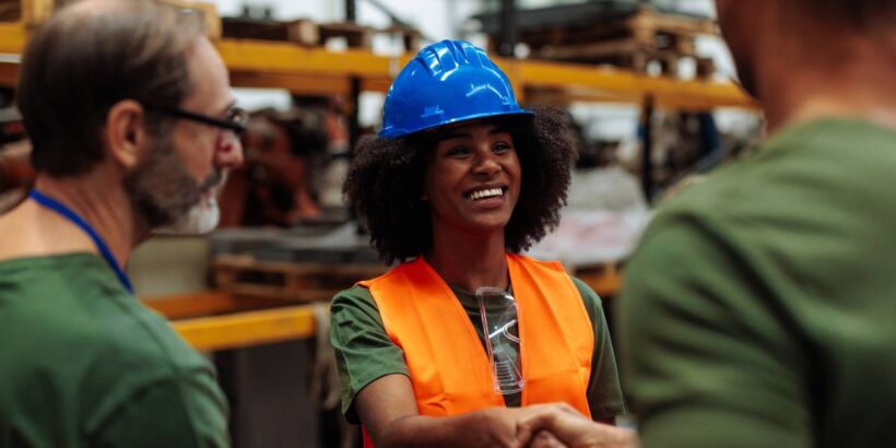 A black woman wearing a blue hard hat and orange vest shakes hands with someone. Another man stands next to them.