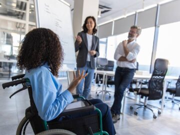 Three people gather around a whiteboard in a business conference room. Two people stand and one sits in a wheelchair.