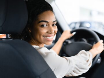 A young Black woman smiling into the backseat of her car. Her hands are on the steering wheel and she's parked.