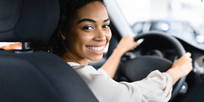 A young Black woman smiling into the backseat of her car. Her hands are on the steering wheel and she's parked.