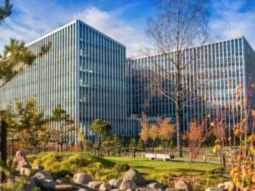 A large office building with a lot of colorful trees in front of it. There is also a water feature surrounded by rocks.