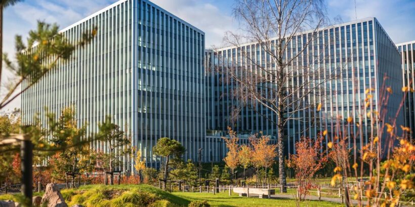 A large office building with a lot of colorful trees in front of it. There is also a water feature surrounded by rocks.