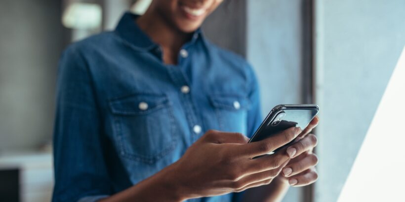 A smiling woman wearing a denim button-up shirt and black glasses uses her thumbs to type on a black mobile device.