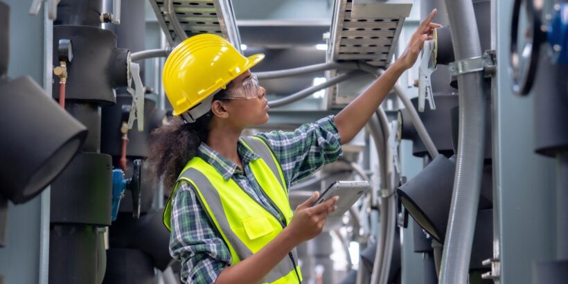 A Black woman in a yellow hard hat and reflective vest pointing at industrial equipment while holding a tablet.