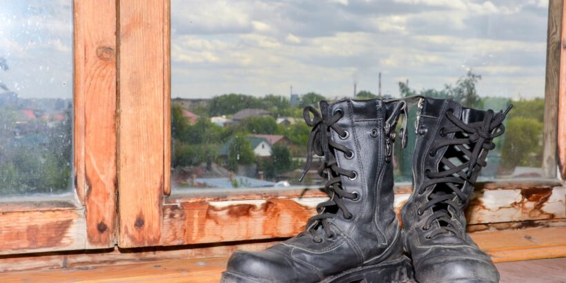 A pair of black military boots is placed near a wooden window frame overlooking a residential neighborhood.