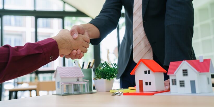 A man wearing a stripe tie and suit shaking hands with another man. On the table between them are miniature model homes.