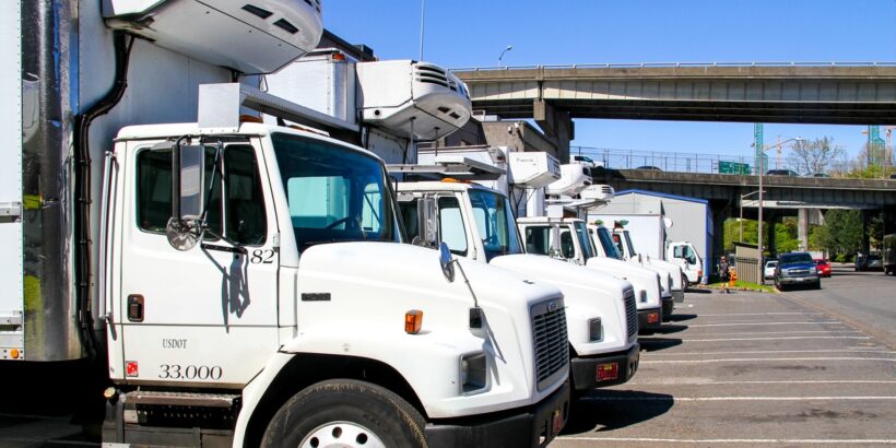 A row of white commercial refrigerator trucks are parked in a lot near an overpass on a sunny afternoon.
