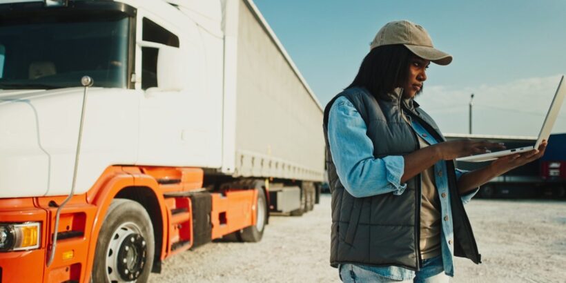 A Black woman wearing a baseball cap uses her laptop outside at a rest stop, standing next to her semi-truck.