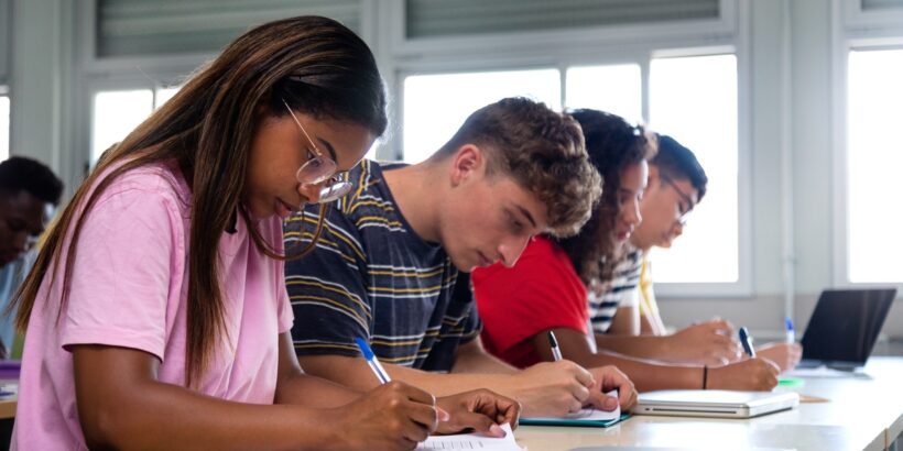 Four students are sitting in a row at a long table. They are writing on paper with blue and black pens.