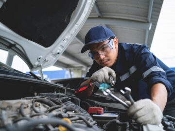 A male auto technician in safety glasses, gloves, and a hat is using tools to repair a white car's engine.