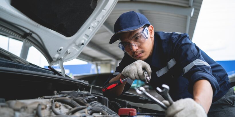 A male auto technician in safety glasses, gloves, and a hat is using tools to repair a white car's engine.