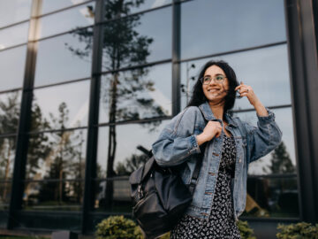 A young woman wearing a jean jacket and carrying a black backpack smiles as she passes in front of a building.