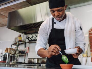 A young, Black chef working in a commercial restaurant kitchen. He is spooning pesto into a small orange bowl.