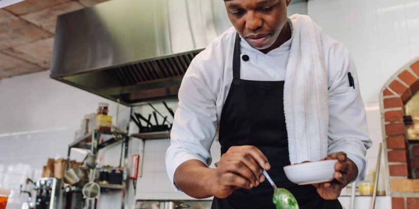 A young, Black chef working in a commercial restaurant kitchen. He is spooning pesto into a small orange bowl.