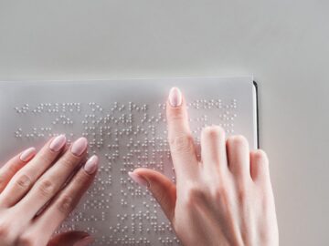 An individual with pink nail polish using their fingers to read the braille text on a white sign with a gray background.