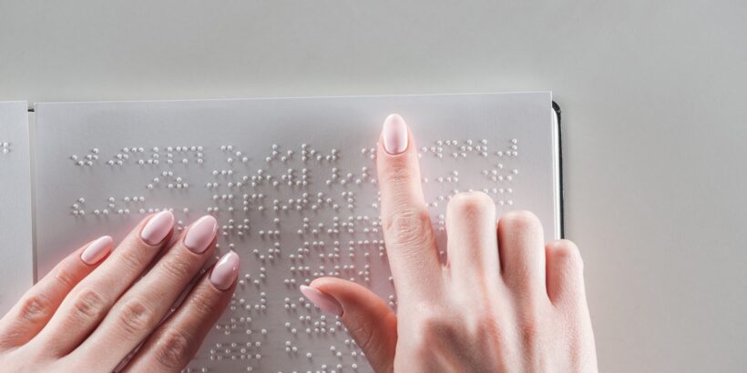 An individual with pink nail polish using their fingers to read the braille text on a white sign with a gray background.