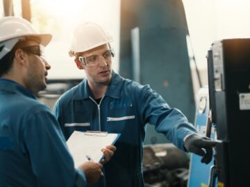 Two male workers in blue uniforms and white hard hats standing in front of a black machine while one man holds a clipboard.