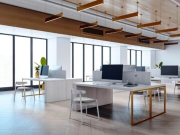 A modern, open-plan workspace with a large wall of windows, hanging light fixtures, and white desks with computers.