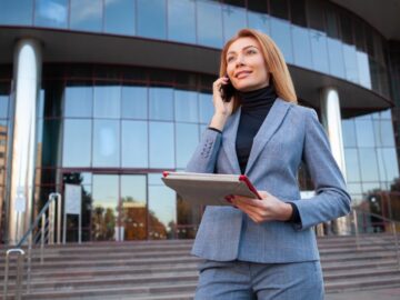 A blonde woman in a blue business suit and black turtleneck using a cell phone in front of a glass-walled building.