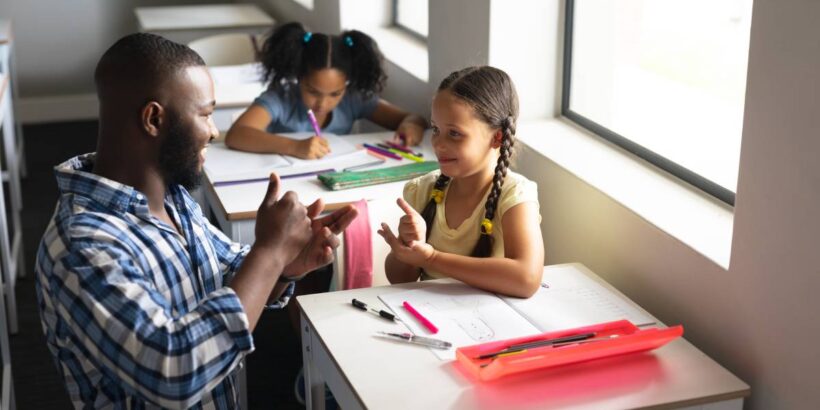 A young girl with braids sitting at her desk at school uses sign language with her smiling Black male teacher.