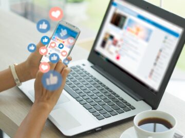 A woman holds her phone in front of a laptop and cup of coffee. Likes and hearts come off the phone screen.