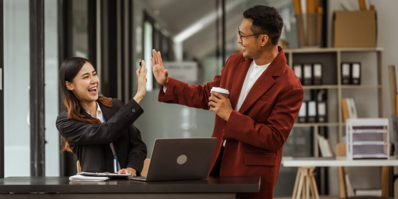 A man and a woman in business attire high five. The man holds a coffee while the woman sits with a laptop.
