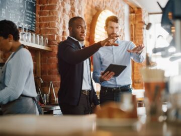 Two professionally dressed men standing behind a coffee counter, gesturing toward the machines. One holds a tablet.