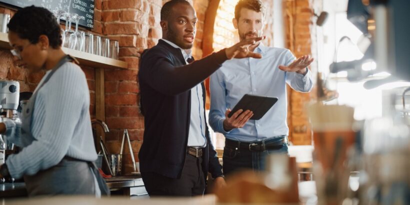 Two professionally dressed men standing behind a coffee counter, gesturing toward the machines. One holds a tablet.