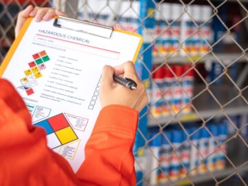 A person in orange protective clothing holding a clipboard with a hazardous chemical checklist near storage shelves.