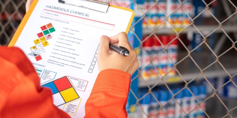 A person in orange protective clothing holding a clipboard with a hazardous chemical checklist near storage shelves.