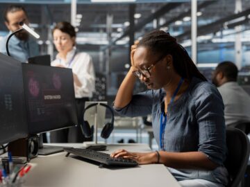A woman looks at the keyboard connected to two monitor computers. She has her head in her hand, frustrated.