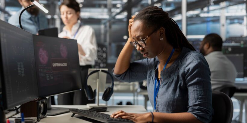 A woman looks at the keyboard connected to two monitor computers. She has her head in her hand, frustrated.