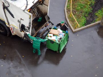 A garbage truck picks up a business's trash from their large green hopper outside. There are two garbagemen sorting.